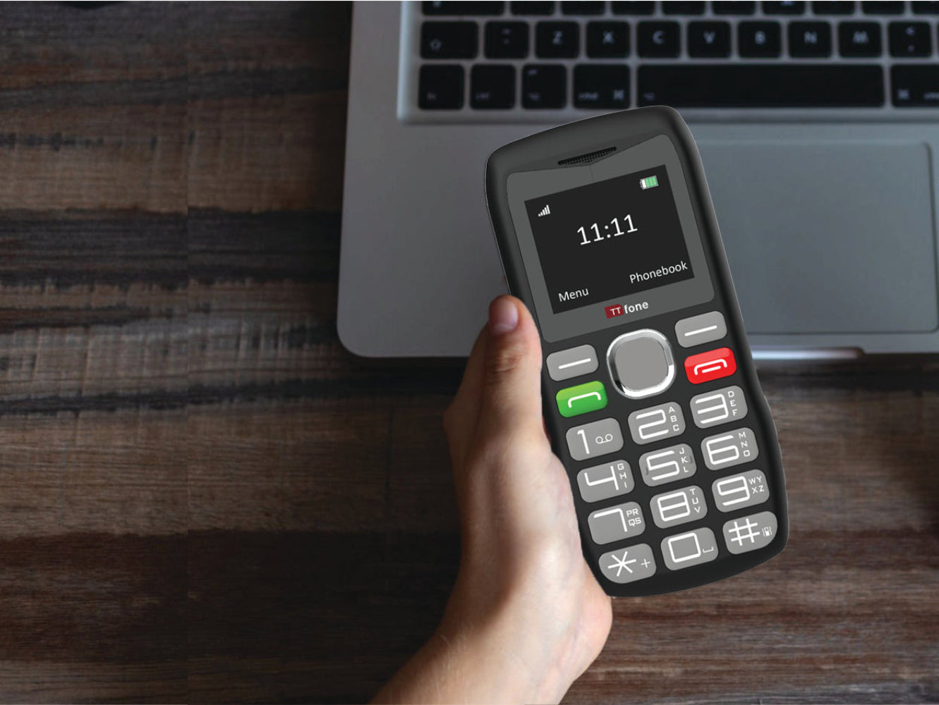 Hand holding a small black phone with a laptop in the background on a wooden surface